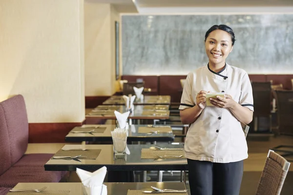 身着白色制服的亚洲年轻女服务员站在桌子旁 在餐厅的记事本上写字时 对着镜头微笑 — 图库照片
