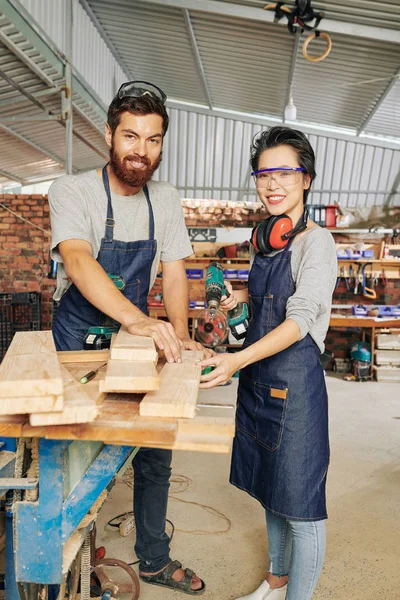 Cheerful multi-ethnic furniture manufacture owners drilling wooden planks on workbench