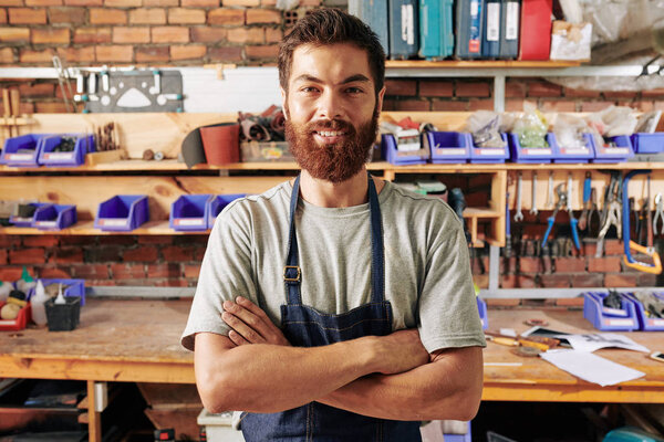 Портрет красивого улыбающегося бородатого плотника, стоящего со скрещенными руками и смотрящего в камеру

