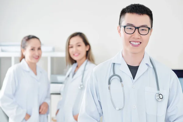 快乐的年轻越南全科医生戴着眼镜对着镜头微笑 他的女同事站在背景 — 图库照片