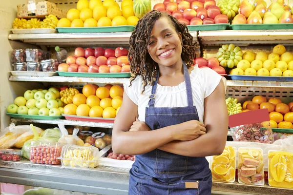 腕を組んで立っているジーンズのアフリカの若い女性の肖像画は 背景に果物や野菜の棚とカメラで微笑んで — ストック写真