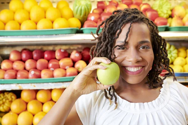 彼女の顔の近くに緑の熟したリンゴを保持し 果物や野菜部門のカメラで微笑むドレッドロックを持つ美しいアフリカの女性の肖像画 — ストック写真