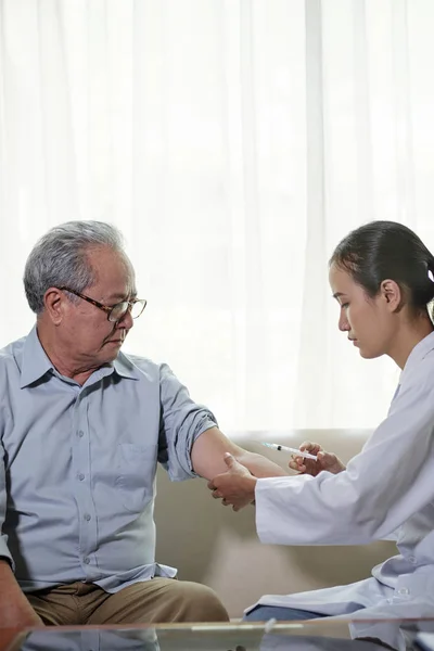 穿着白色外套的年轻护士坐在一个坐到家里的老年病人手中打针 — 图库照片