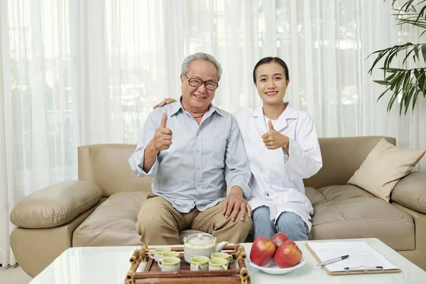 快乐老年病人和他的护士坐在一张大拇指和微笑的相机 而在家里喝茶的肖像 — 图库照片
