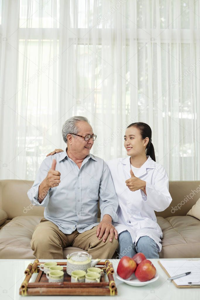 Happy senior patient and young nurse showing thumbs up and looking at each other while sitting on sofa and drinking tea nurse coming to visit him