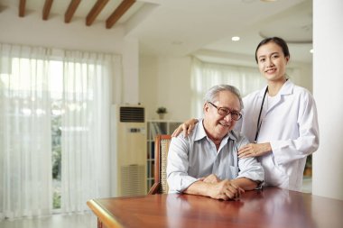Masada oturan ve hastanede yanında duran beyaz önlüklü kadın doktoruyla kameraya gülümseyen kıdemli hastanın portresi