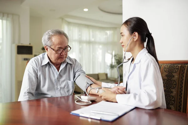 アジアの若い看護師は 高齢の男性と一緒にテーブルに座って 自宅で彼女の訪問中に彼の血圧を調べる — ストック写真