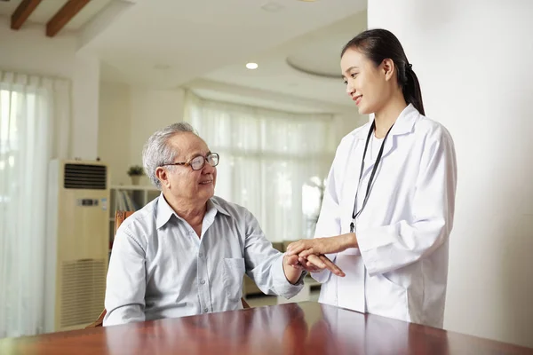 亚洲女医生穿着白色外套 握着她的老年病人的手 微笑着 而他坐在桌旁 在医院与她交谈 — 图库照片