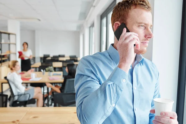 穿着蓝色衬衫的年轻商人用手机交谈 透过窗户看着在办公室后台工作的商人 — 图库照片
