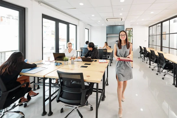 Young Asian businesswoman or secretary with folders walking along the office corridor with other business people working at their workplaces