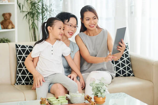 デジタルタブレットを持ち 自宅のソファに座っている間 彼女の家族に何かを示す笑顔のアジアの女性 — ストック写真