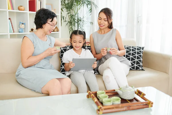 家庭一代放松在家里的母亲和祖母喝茶从小杯子 而小女儿坐在和使用数字平板电脑在家里 — 图库照片