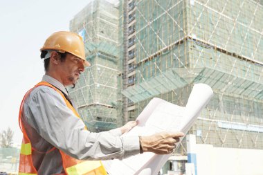 Hardhat ve yansıtıcı giyim genç mühendis inşaat halindeki binanın karşısında açık havada dururken planı inceleyerek