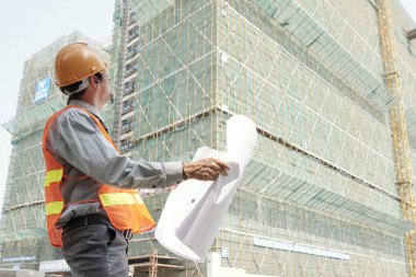 İnşaat aşamasındaki yüksek binaya bakan hardhat ve yansıtıcı yelek giyen mühendis, dışarıda planla ayakta dururken ve çizimleri kontrol ederken