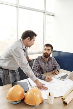 Masada oturan iki genç işadamı, planlar ve dizüstü bilgisayarla çalışarak ofiste birlikte yeni inşaat projesini tartışıyorlar.