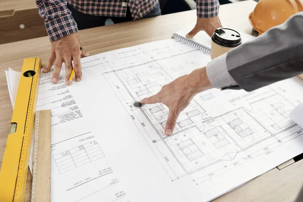 Close-up of business people standing near the table pointing at construction blueprint examining and discussing it together during meeting at office