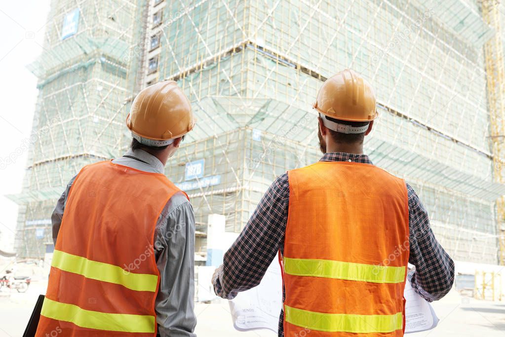 Rear view of two construction workers in hardhats and reflective vests holding blueprint and looking together at high building under construction outdoors