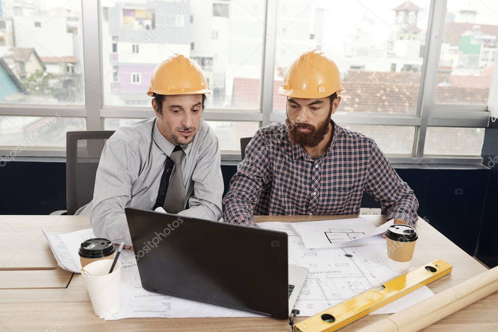 Two business partners in hardhats sitting at the table and working on laptop computer during a meeting at office