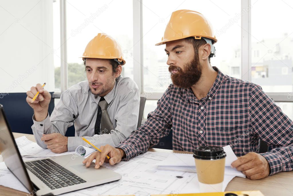 Two architects wearing hardhats sitting at the table using laptop computer and discussing working moments together at office