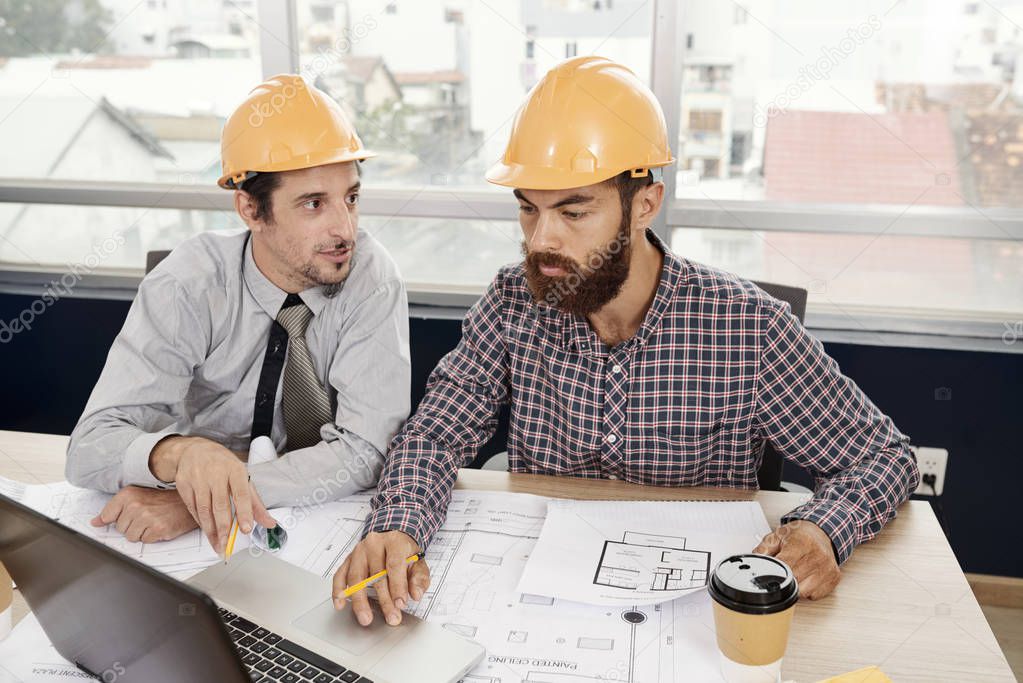 Two architects in hardhats sitting at the table with blueprint and using laptop computer in work at business meeting at office