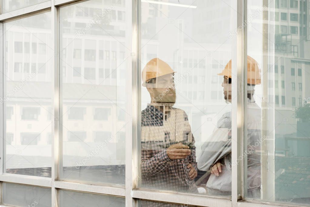 Two architects in hardhats talking to each other while standing behind the glass wall at modern office