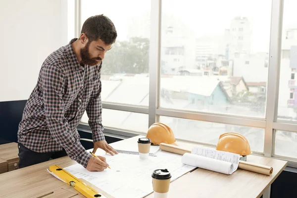職場の近くに立ち オフィスで鉛筆で建設の青写真を描くカジュアルな服装の若いひげエンジニア — ストック写真