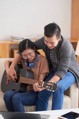 Asyalı mutlu çift kanepede oturan ve evde boş zamanlarında birlikte gitar çalmayı öğrenme