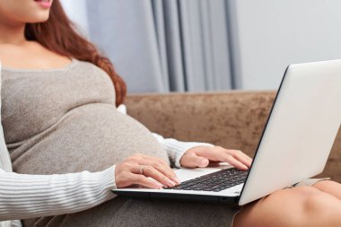 Evde kanepede oturan ve internette hamilelik hakkında bilgi arayan genç kadının kırpılmış görüntüsü