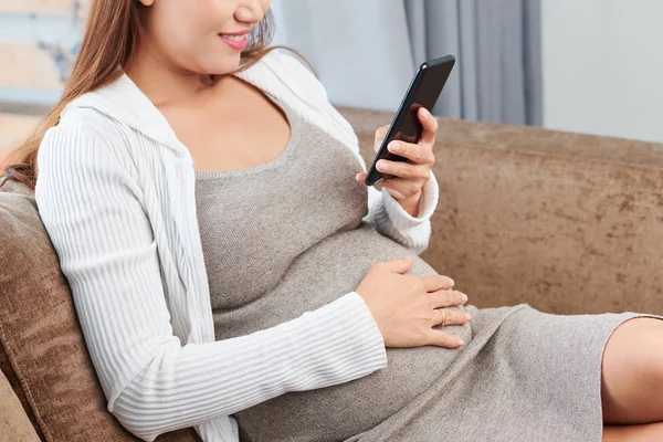 微笑的年轻孕妇触摸她的肚子时 阅读文章和短信在智能手机 — 图库照片