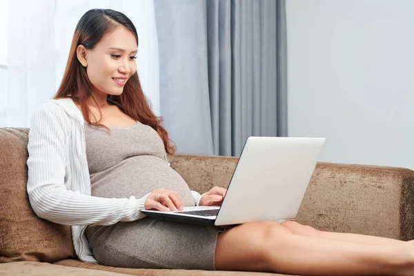 漂亮的年轻的亚洲孕妇穿着羊毛连衣裙坐在一个坐 在家里的笔记本电脑工作 — 图库照片