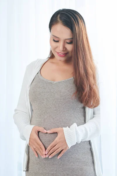 相当积极的年轻越南孕妇看着她的肚子 使心脏形状用手指 — 图库照片