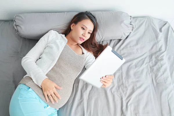 怀孕的亚洲年轻女子躺在床上 在平板电脑上阅读文章 — 图库照片