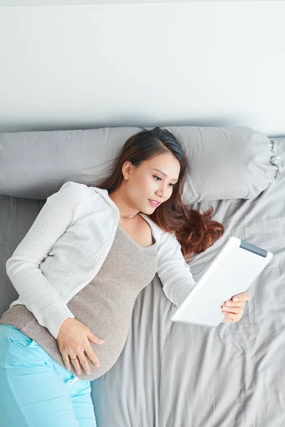 Positive smiling young Asian pregnant woman resting on bed and enjoying reading e-book on weekend