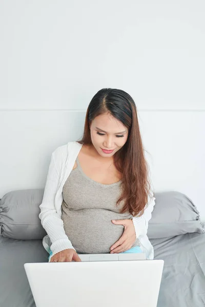 美丽的年轻女子期待婴儿触摸肚子和阅读有关怀孕的文章在线笔记本电脑 — 图库照片