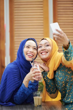 Kokteyllerle kafe masasında otururken akıllı telefonda selfie pozu veren türbanlı neşeli genç Müslüman kadınlar