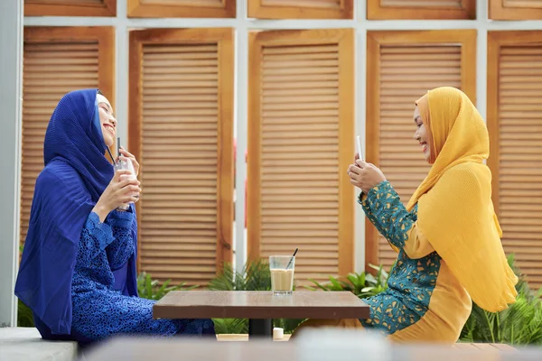 Cheerful young muslim woman in hijab taking photo of her friend with sweet milky cocktail on smartphone