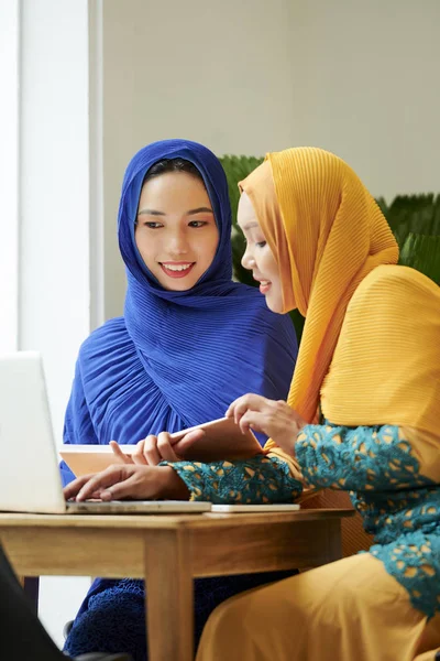 Smiling muslim women in traditional dresses and hijabs reading book and watching webinar on laptop