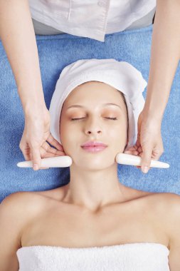 Cosmetologist rahatlatıcı genç Kafkas kadının yüzüne masaj yaparken taş çubuklar kullanarak, yukarıdan görünümü