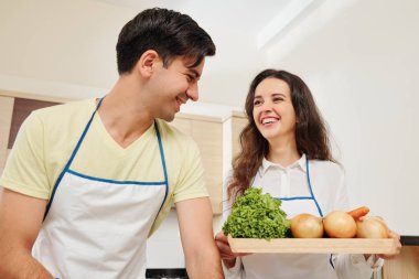 Üzerinde taze sebze olan büyük tahta tahta taşıyan karısına bakan gülümseyen genç adam