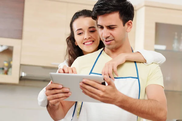 年轻的白种夫妇在围裙站在厨房和阅读数字平板电脑的菜食谱 — 图库照片