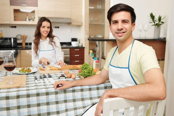 夕食のテーブルに座ってカメラを見ているエプロンのハンサムな若者の肖像画 彼の妻は背景にパンを切る — ストック写真