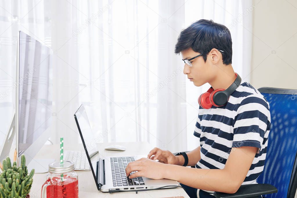 Serious mixed-race boy in glasses with headphones around his neck working on laptop in his room