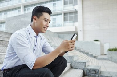 Gülümseyen genç Asyalı işadamı merdivenlerde oturan ve cep telefonu metin mesajları okuma 