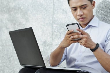 Ciddi Vietnamlı yazılım geliştiricisi açık dizüstü bilgisayar ile açık havada oturan ve telefonunda haberci kontrol