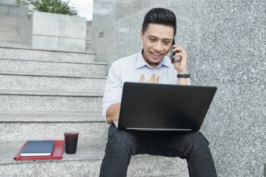 Neşeli genç Asya üniversite öğrencisi merdivenlerde oturan, telefon arama ve dizüstü bilgisayar ekranında e-postaları kontrol