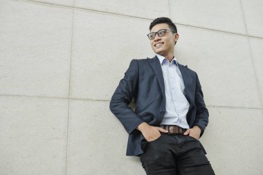 Neşeli genç yakışıklı Vietnamlı girişimci duvara yaslanmış ve uzağa bakıyor