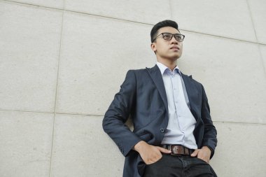 Gözlüklü kendinden emin genç Vietnamlı işadamı bina duvarında dururken ellerini ceplerine koyarak 