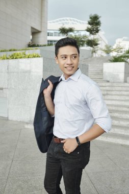 Yakışıklı gülümseyen genç Asyalı girişimci omzunda ceket ile açık havada ayakta