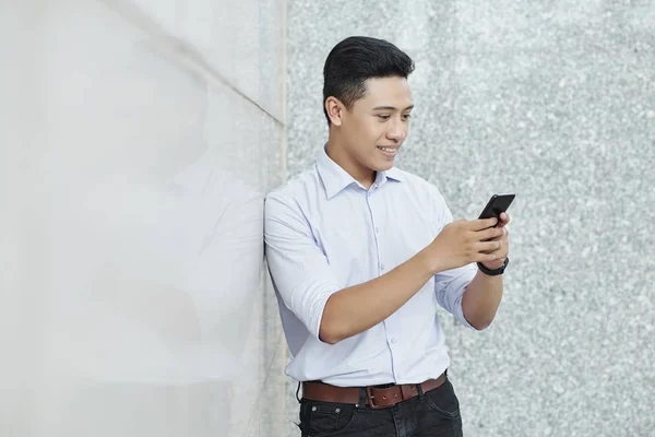 携帯電話でテキストメッセージを送るとき 壁に寄りかかっている若いアジアの起業家を笑顔 — ストック写真