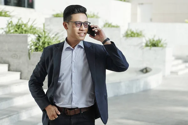 Portrait of attractive successful young entrepreneur in glasses standing outdoors with hand in pocket and making phone call